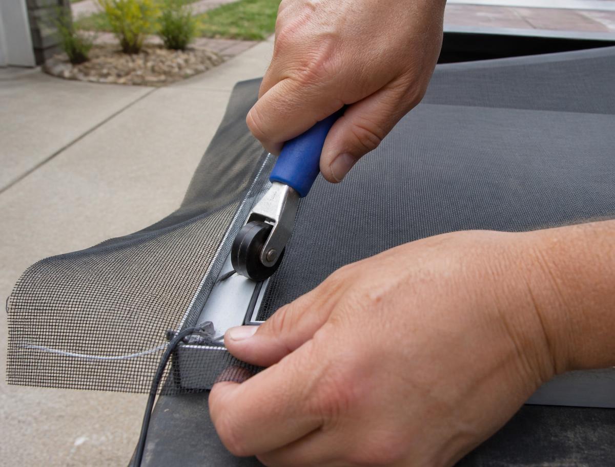 Hands rolling new charcoal fiberglass mesh into an aluminum window screen frame